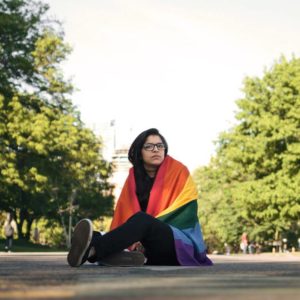 a light brown skinned person is sitting on the ground; she has long hair and is wearing glasses and has a rainbow flag wrapped around her. The background has trees on both sides of her.