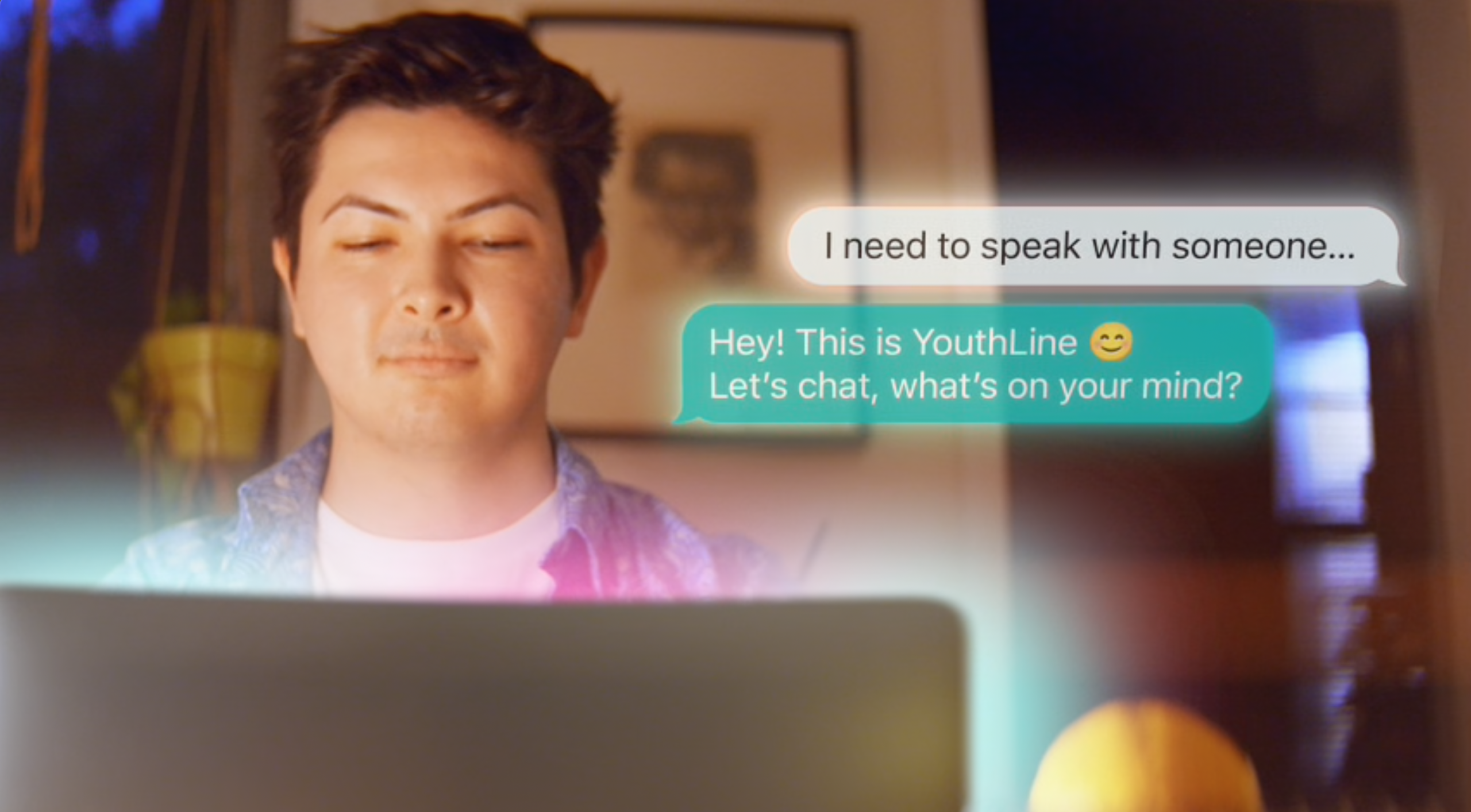 A screenshot from the LGBT YouthLine promotional video "Someone Like Me: Peer Support and LGBT YouthLIne". A person sits in a room looking at a laptop. Text bubbles float in the air. One reads "I need to speak with someone". The other reads "Hey! This is YouthLine. Let's chat. What's on your mind?"